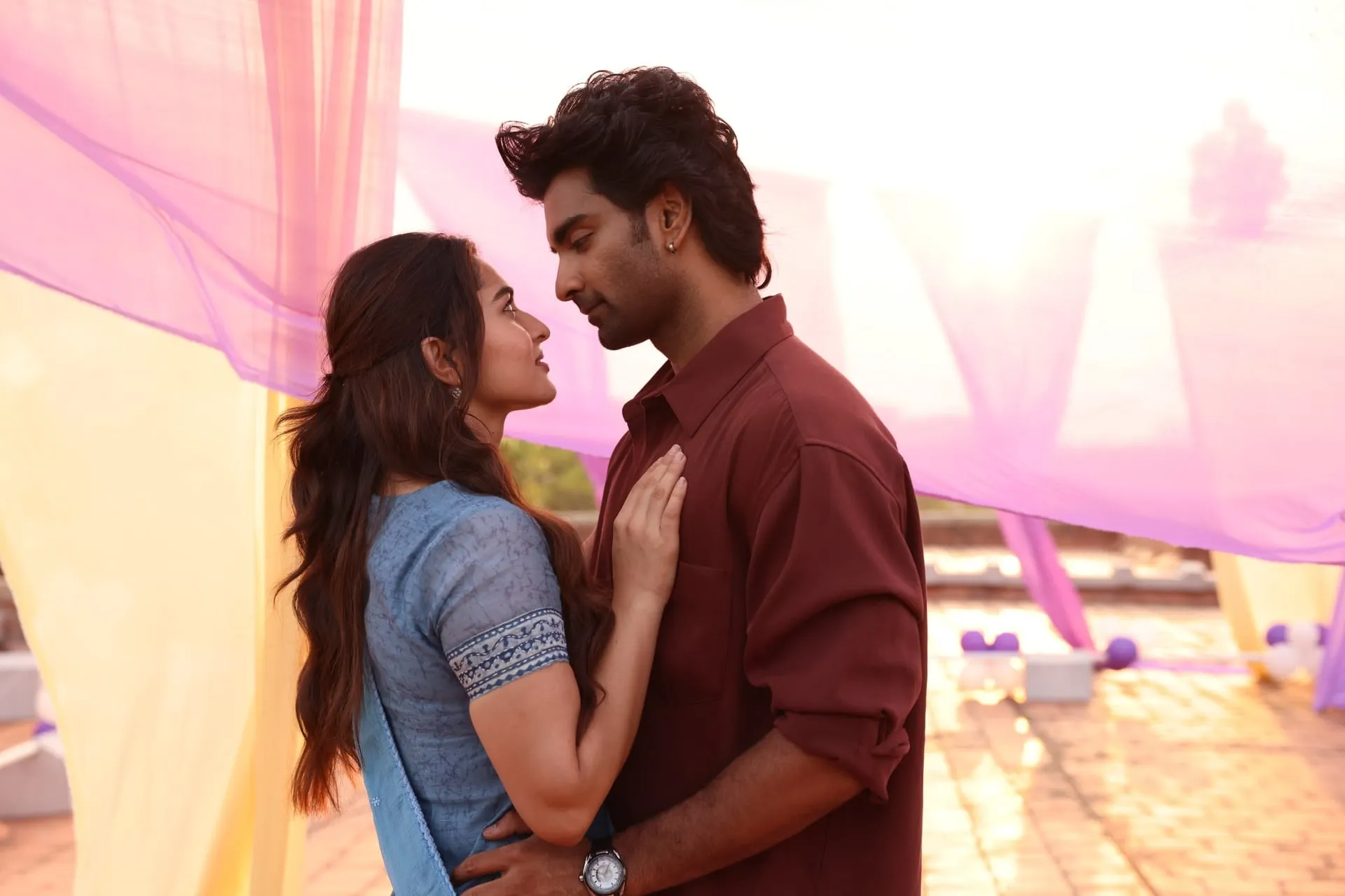 Idhaya and Sam stand close together under fluttering pastel drapes in a warm rooftop scene from Idhayam Murali