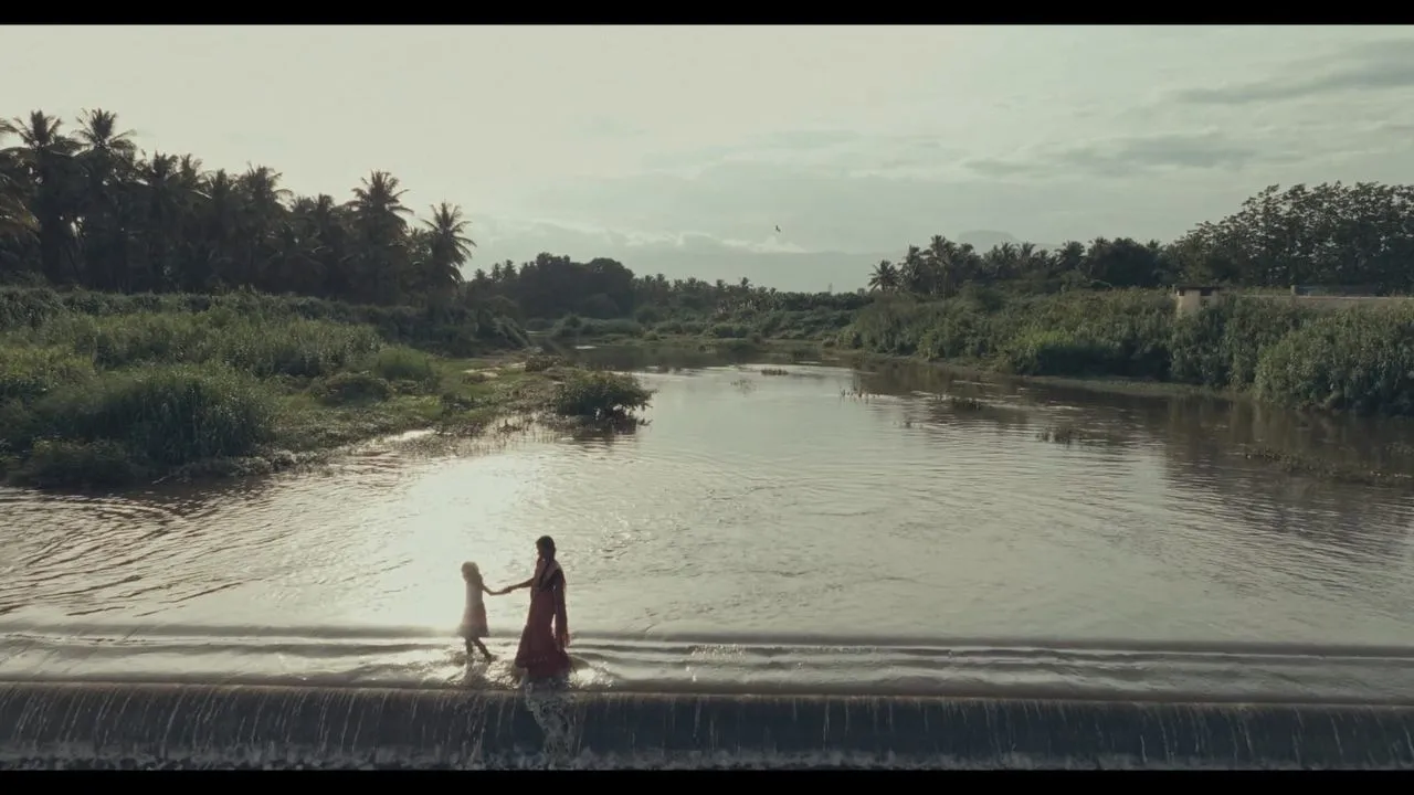 A still from Mayilaa showing two figures crossing a river against a lush Tamil Nadu landscape