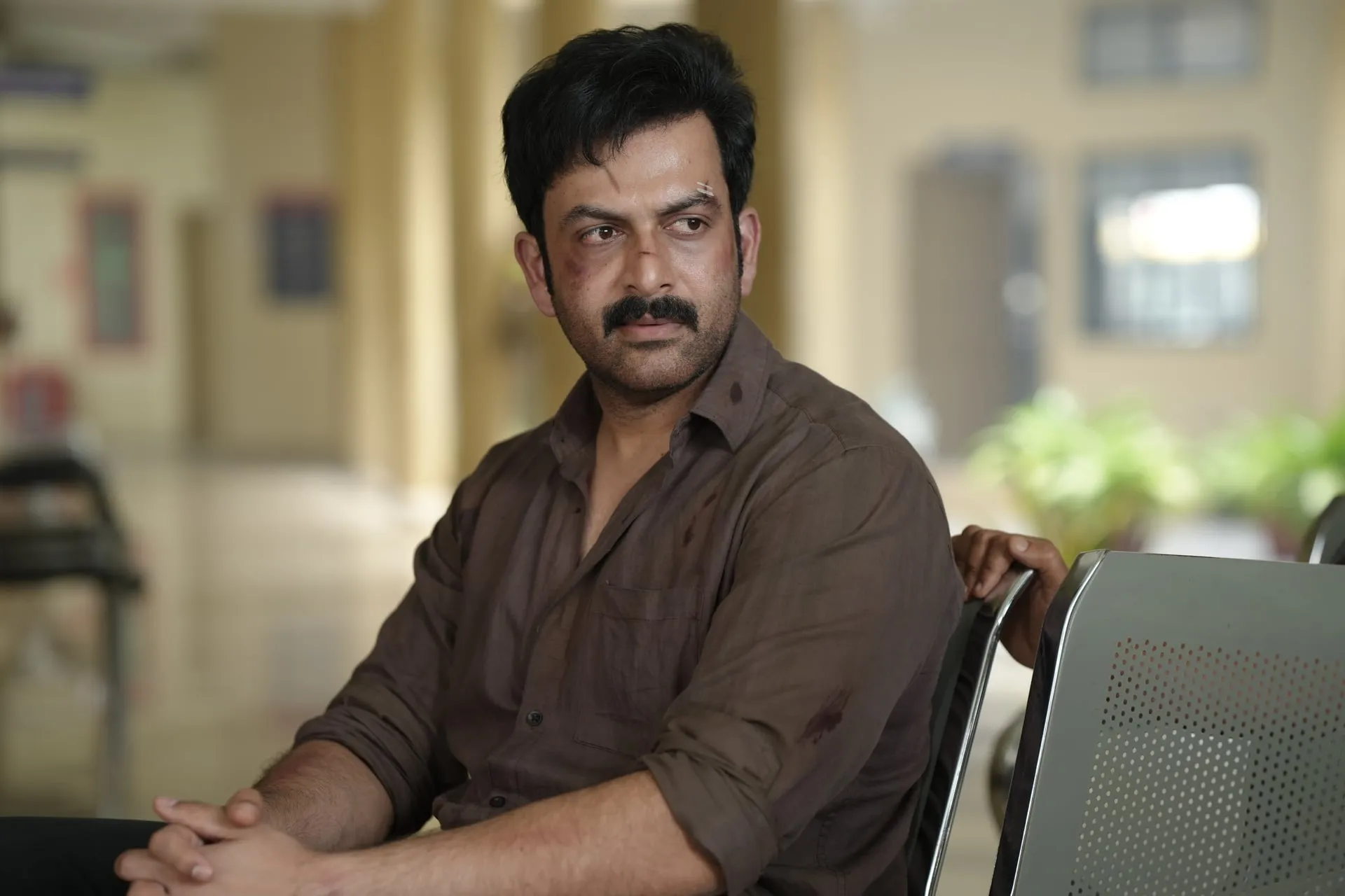 Rajeevan, bruised and weary in a brown shirt, sits alone on a bench in a government building corridor