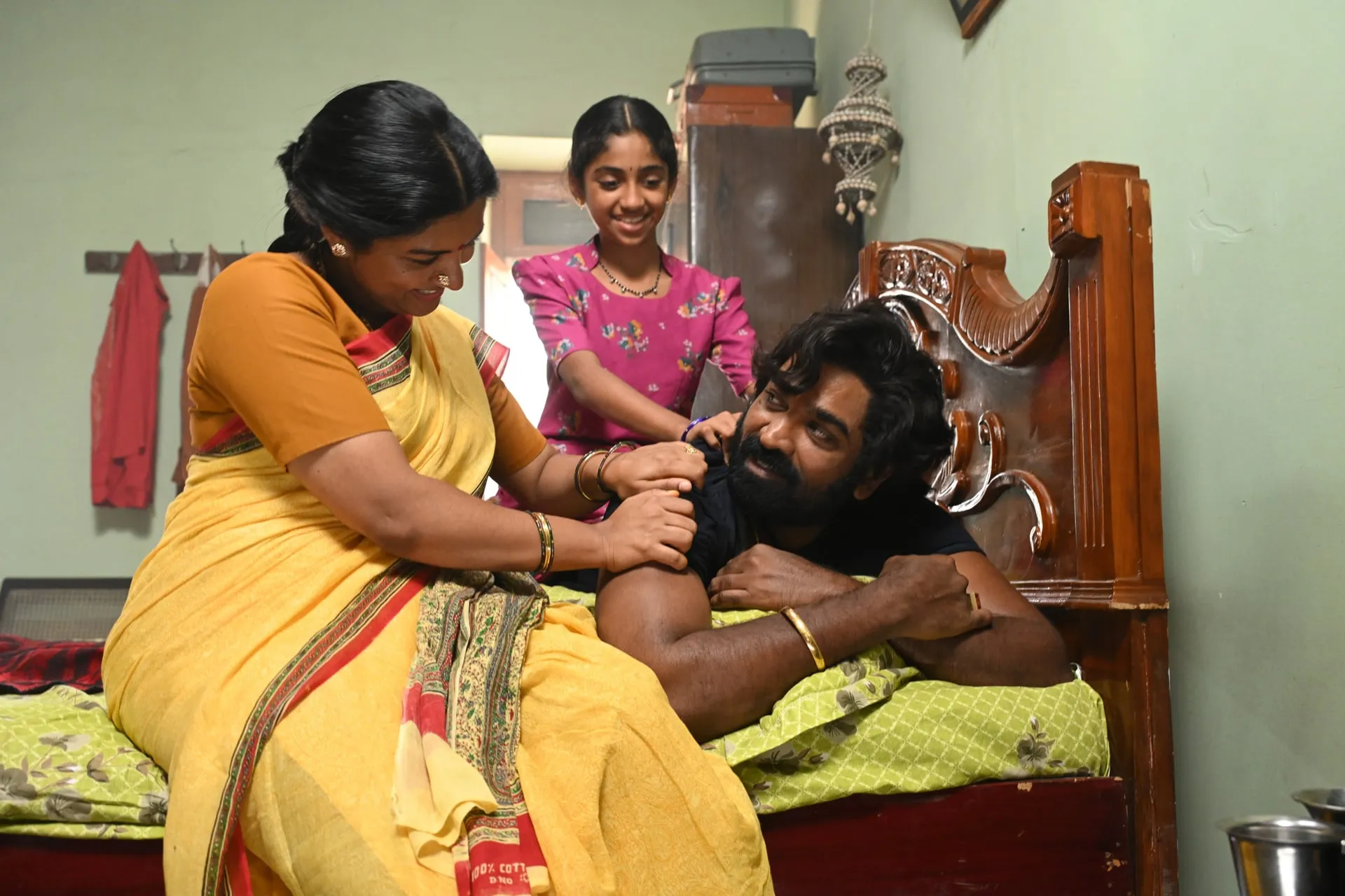 Vijay Sethupathi lies on a bed as Lijomol Jose massages his shoulders and a young girl leans over him in a 1980s bedroom in Baththa