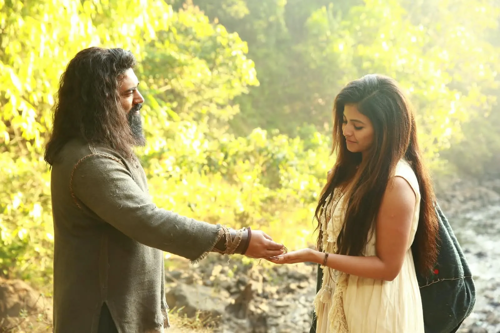Nivin Pauly, long-haired and bearded in a sackcloth tunic, places something in Anjali's open palm in a sunlit forest in Yezhu Kadal Yezhu Malai