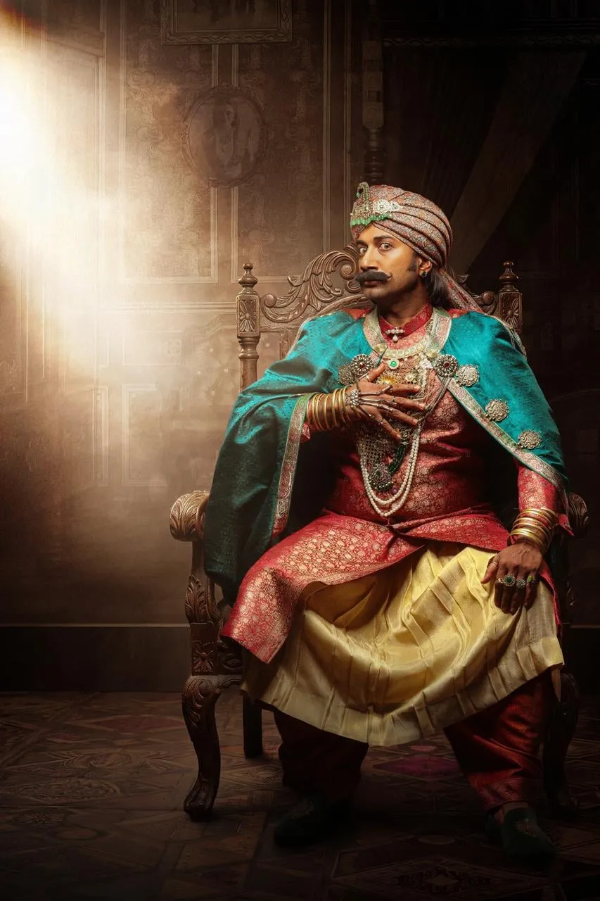 Satya Dev as Ramappa Rao Bahadur on a carved wooden throne in a red brocade kurta, teal cape and jewelled turban, one ringed hand pressed to his chest