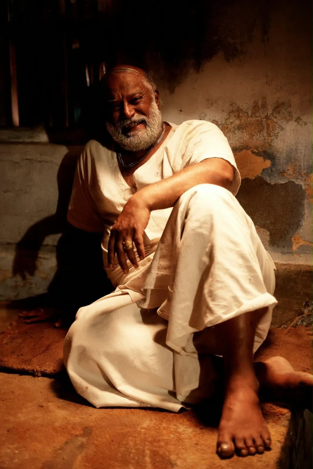 Vettai Muthukumar seated barefoot on a mud floor in a white veshti