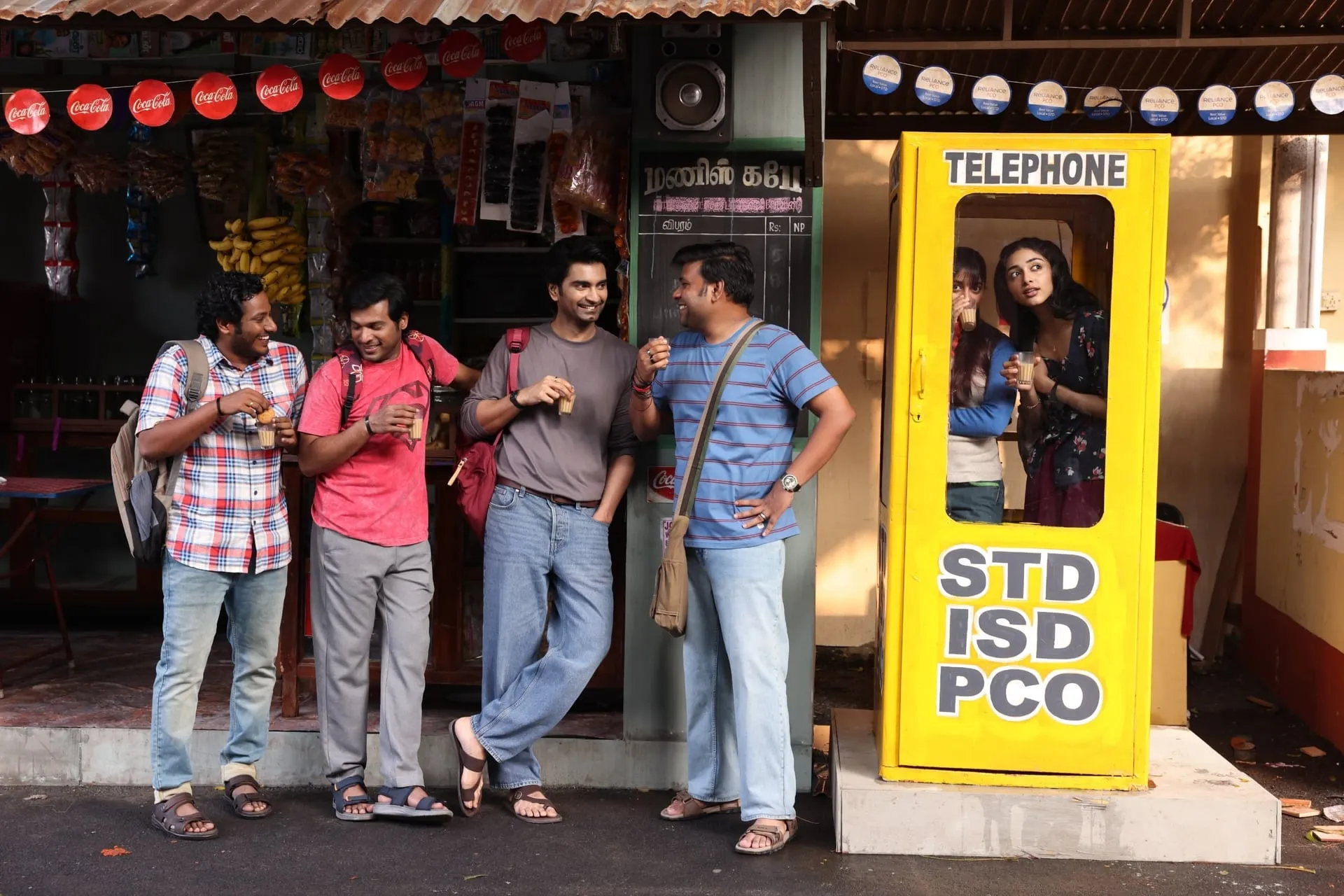 Idhaya and his friends drink tea outside a roadside shop with an old yellow STD ISD PCO telephone booth