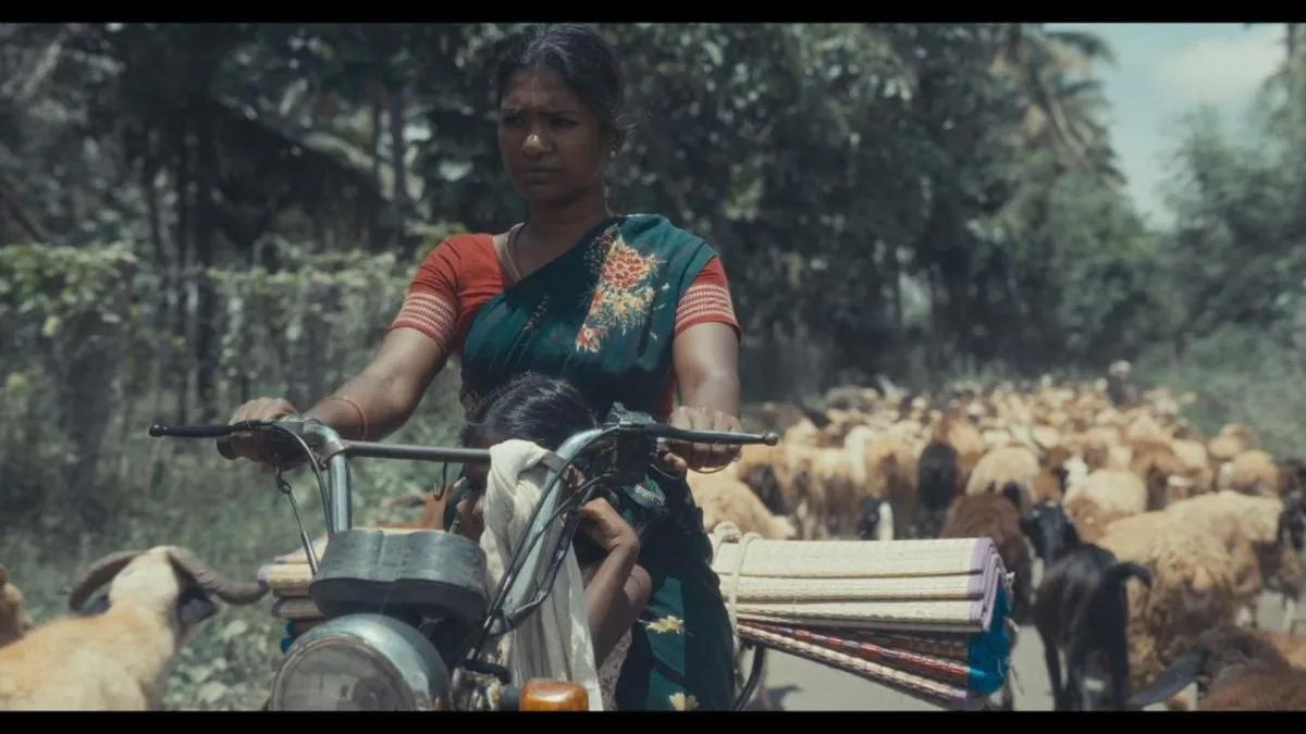 P. Melody Dorcas riding a bicycle with a child, a flock of sheep in the background, in a still from Mayilaa