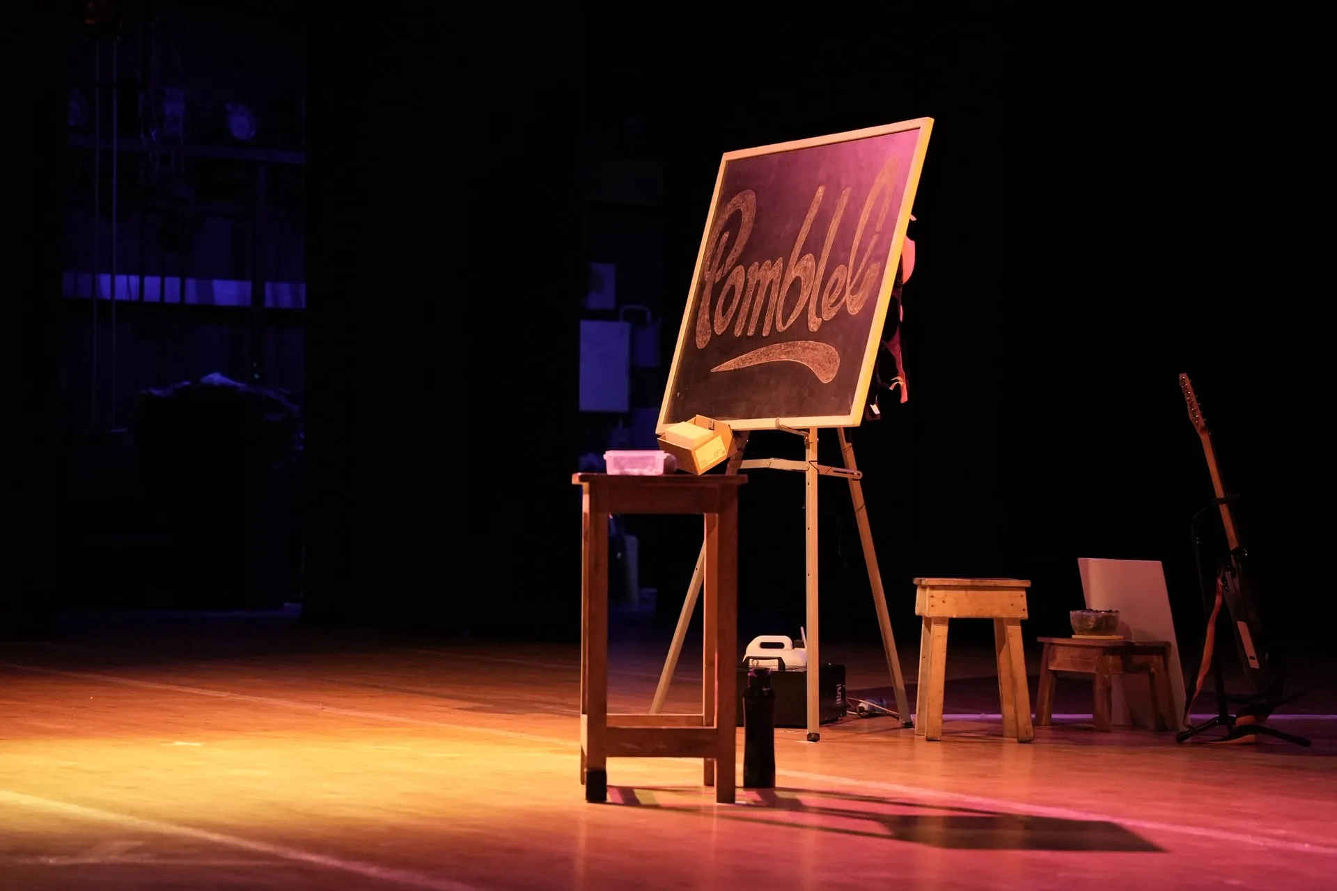 The Pomblel chalkboard standing on the Music Academy stage, a guitar waiting at the edge of the light