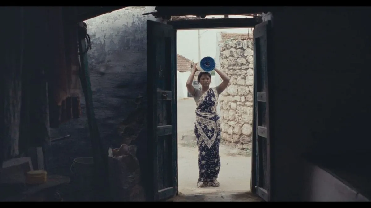 A woman framed in a doorway carrying a blue rolled mat on her head in a still from Mayilaa