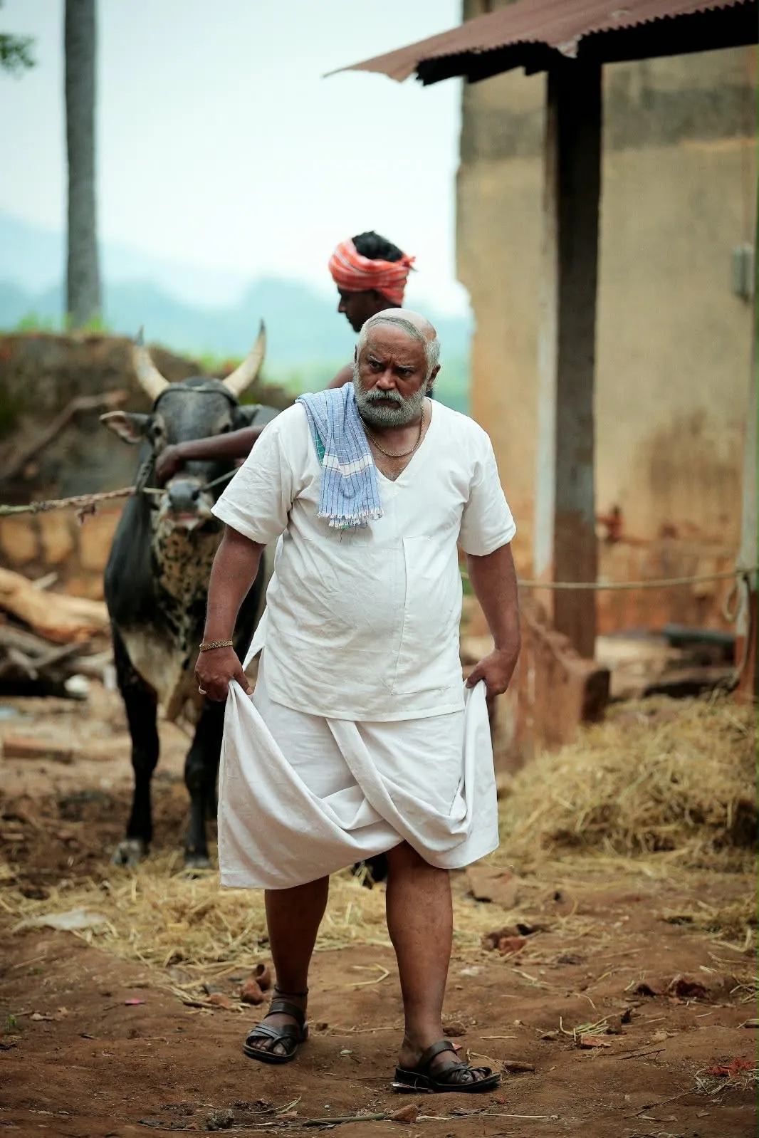 Vettai Muthukumar walking past a bull in a village courtyard, a towel over his shoulder