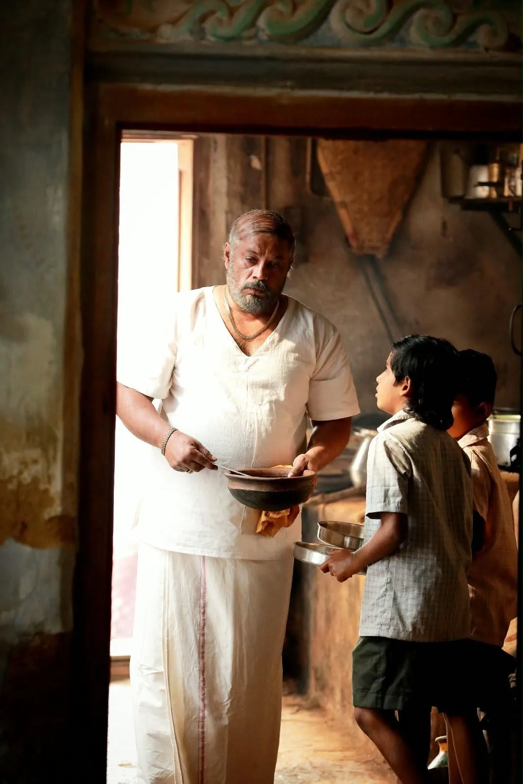 Vettai Muthukumar ladling food in a kitchen doorway as two boys wait