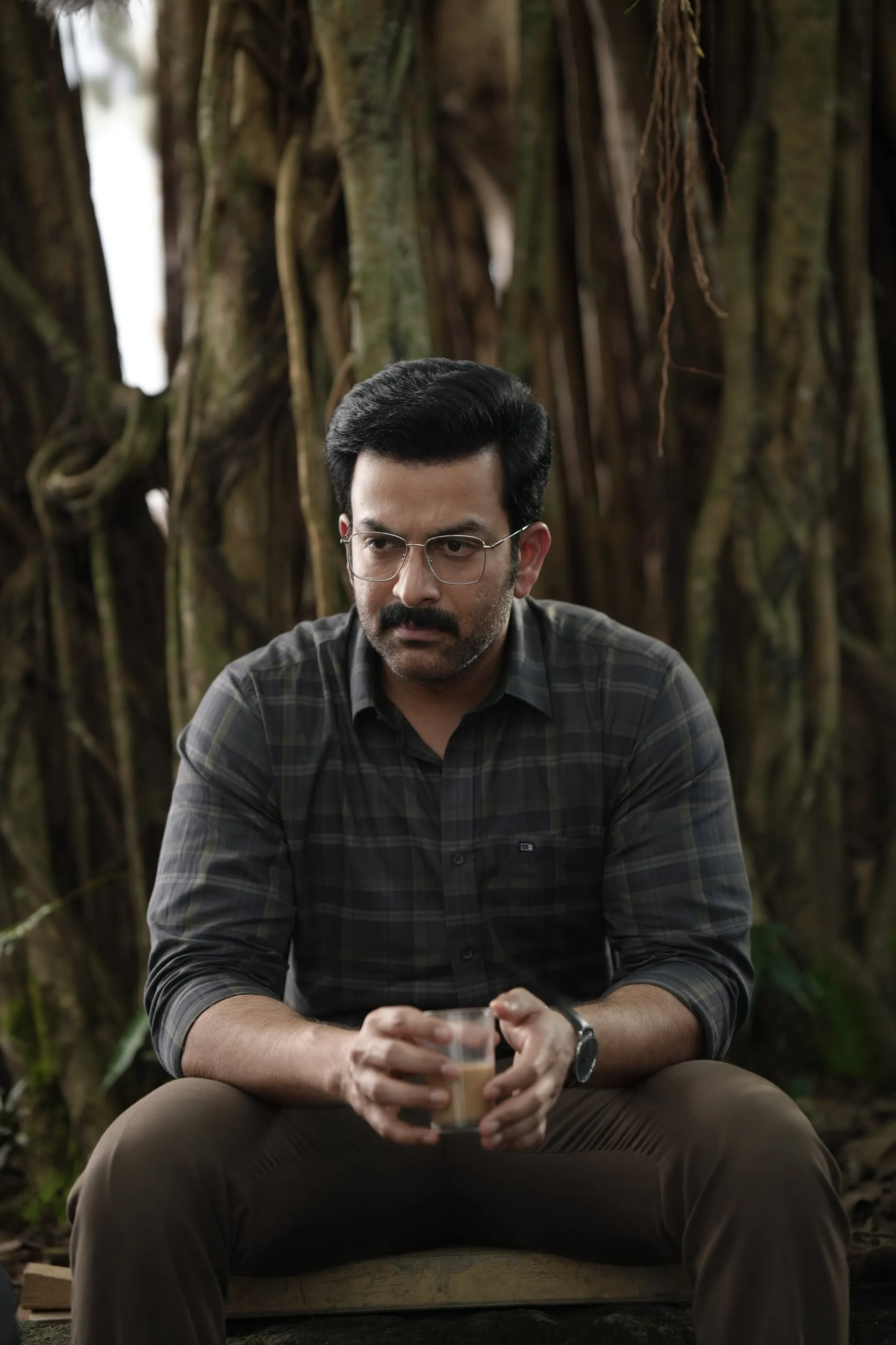 Rajeevan sits alone under a banyan tree, holding a glass of tea, lost in thought