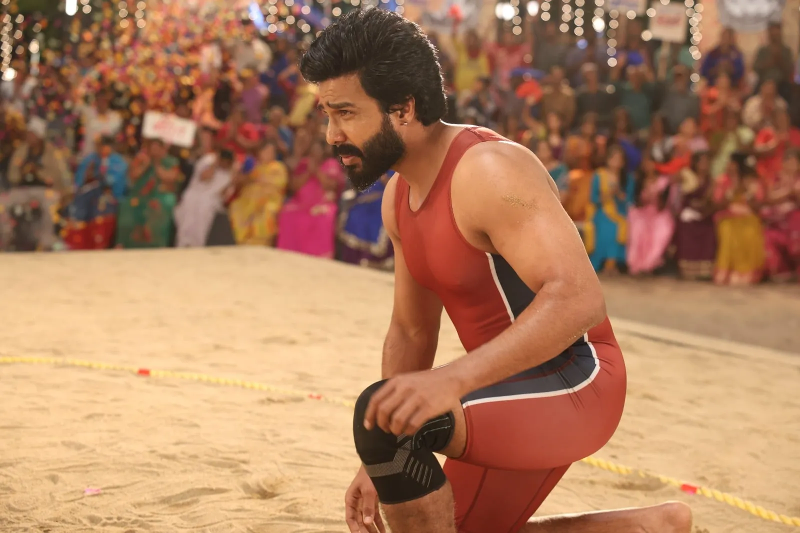 Vishnuu Vishal in a red wrestling singlet kneeling on the sand of a crowded village arena in Gatta Kusthi 2